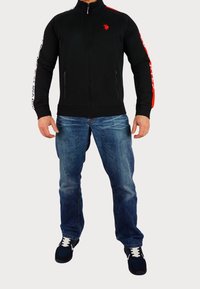 Giacca nera con zip, dettagli rossi e cintura con logo bianco sulle maniche, abbinata a jeans blu e sneakers scure. Sfondo neutro e solido.