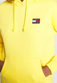 Felpa gialla con tasca frontale, cappuccio con cordino e un piccolo tag nero e colorato con la scritta "TOMMY JEANS." Materiale in cotone morbido.