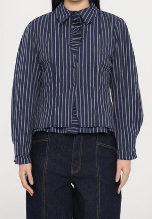 Kvinde iført en mørkeblå bluse med lange ærmer, hvide lodrette striber og flæsedetalje, kombineret med mørkeblå højtaljede jeans.