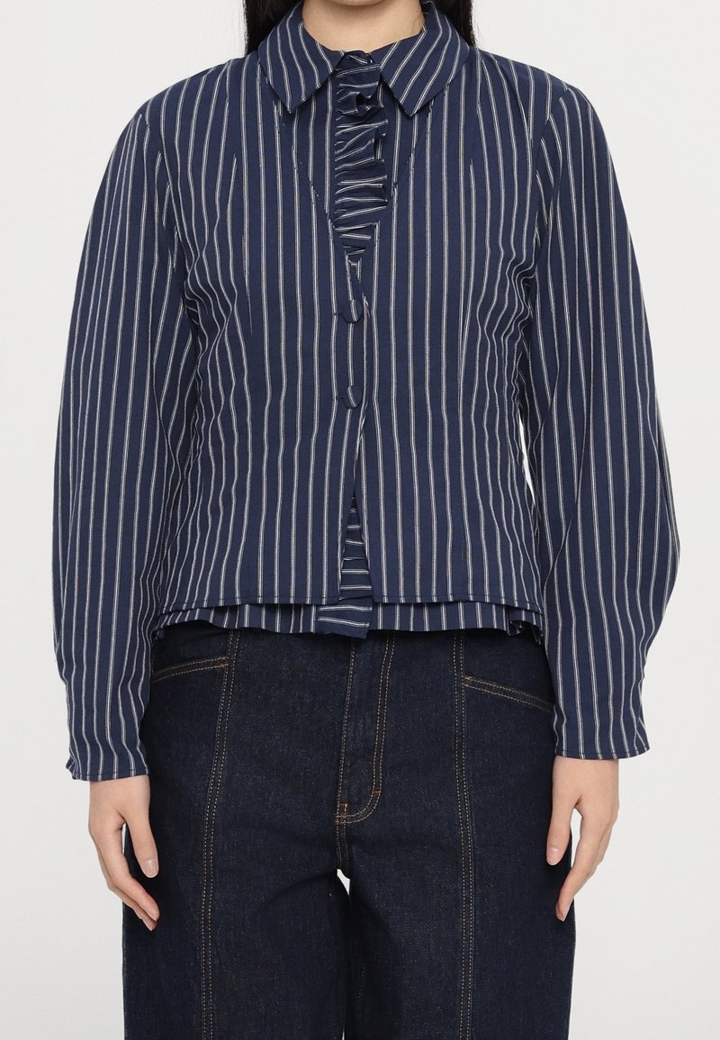 Femme portant un chemisier bleu marine à manches longues avec des rayures verticales blanches et un détail volanté, associé à un jean taille haute bleu foncé.