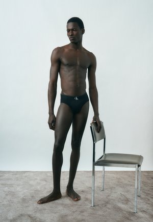 Mužský model v čiernych slipoch Calvin Klein stojí bosý na koberci, jednou rukou sa opiera o sivú čalúnenú stoličku s kovovými nohami.