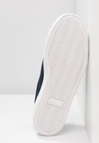 Semelle de chaussure blanche avec un motif de bande de roulement en zigzag et le logo "NO NAME" embossé au centre, inclinée contre une surface blanche.