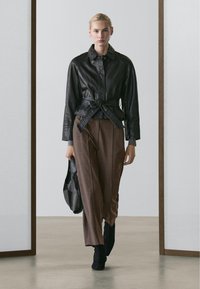 Kvinde går indendørs iført en sort læderjakke bundet i taljen, brune vidde bukser, sorte støvler og bærende en sort taske.