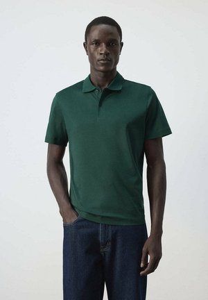 Uomo che indossa una polo verde scuro a maniche corte e jeans blu scuro, in piedi davanti a uno sfondo chiaro.