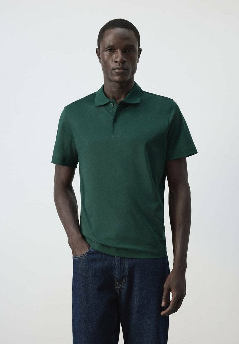 Homme portant un polo uni vert foncé à manches courtes et un jean bleu foncé, debout devant un fond clair.