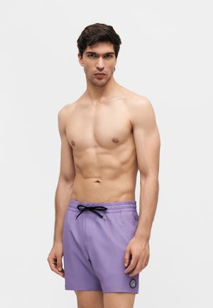 Shirtless man wearing light purple swim shorts with black drawstring and circular patch, standing against a plain white background.