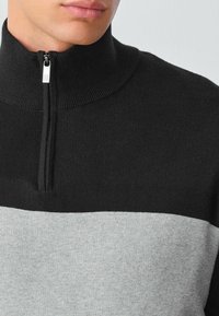 Černý a šedý svetr na zip s vysokým límcem, ukazující barevně blokovaný design. Tkanina vypadá texturovaně a pleteně. Viditelné detaily se stříbrným zipem.