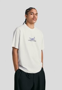 White cotton t-shirt featuring a relaxed fit, round neckline, and purple printed text. Paired with black relaxed-fit trousers.