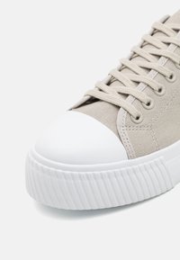 Baskets en toile beige avec un bout rond en caoutchouc blanc, semelle blanche texturée et lacets beiges assortis.