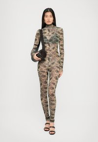 Mujer vistiendo un body transparente con un patrón floral, sosteniendo un bolso negro redondo y usando sandalias de tacón negras, en un fondo simple.
