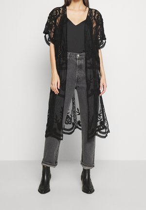 Donna che indossa un cardigan nero in pizzo trasparente sopra una maglietta nera, jeans grigi a vita alta e stivaletti neri, in piedi davanti a uno sfondo bianco.