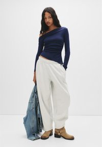 Marin off-the-shoulder topp, åtsittande kropp; ljusgrå baggy sweatpants; bruna klackboots vid anklarna; blå denimjacka över armen.