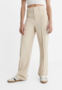 Beżowe spodnie o szerokich nogawkach z wysokim stanem, zapinane na guziki, z delikatnymi plisami z przodu. Do tego białe trampki z czarnym akcentem.