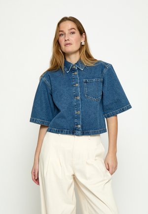 Kobieta o długich jasnobrązowych włosach, ubrana w niebieską jeansową kurtkę z krótkim rękawem i guzikami oraz białe, szerokie spodnie z wysokim stanem, stojąca.