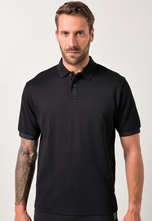 Mann mit Bart und tätowiertem linken Arm, der ein schlichtes schwarzes Poloshirt mit kurzem Arm trägt und vor einem hellen Hintergrund steht.