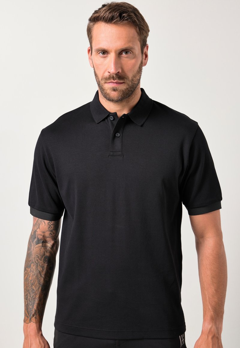Hombre con barba y brazo izquierdo tatuado, vistiendo una camiseta polo negra de manga corta sin estampado, de pie contra un fondo claro.