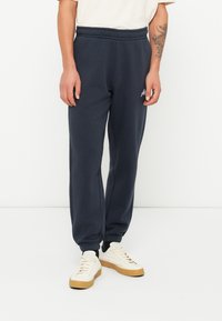 Mørkeblå joggebukser med snor i midjen, ribbede mansjetter og en liten logo på benet. Laget av myk, glatt stoff.