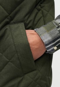 Πράσινο μπουφάν με ραφή σε σχήμα καπιτονέ και τσέπη με ανάγλυφο λογότυπο "Barbour", φορεμένο πάνω από γκρι και πράσινο καρό πουκάμισο με κουμπωτά μανίκια.