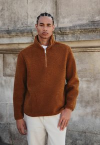 Brauner Fleece-Pullover mit hohem Kragen und möglichem Reißverschluss, mit einer weichen, strukturierten Oberfläche. Über einem weißen Hemd getragen, kombiniert mit hellen Hosen.