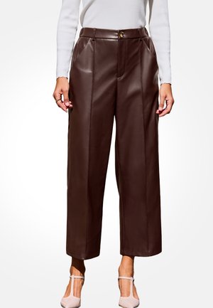 Vrouw draagt een donkerbruine leren broek met hoge taille en wijde pijpen, voorzien van voorplooien, gecombineerd met een witte top met lange mouwen en lichtroze pumps.