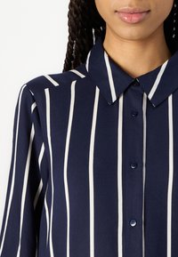Camicia blu navy con strisce verticali bianche, dotata di colletto, chiusura a bottoni e tessuto liscio.