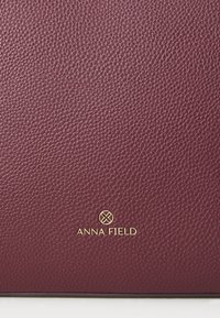 Bordeaux gekorreld materiaal met een gepolijste afwerking. Heeft een gouden logo "ANNA FIELD" gecentreerd aan de onderkant voor branding.
