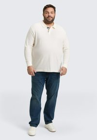 Helle beige Langarm-Polohemd mit einem Kragen mit zwei Knöpfen, kombiniert mit dunkelblauen Jeans und weißen Turnschuhen vor einem neutralen Hintergrund.