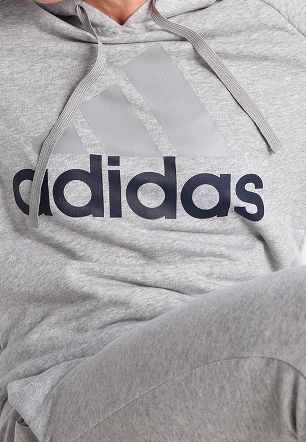 Personne portant un sweat à capuche Adidas gris clair et un pantalon de survêtement assorti, avec un grand logo Adidas sur la poitrine.