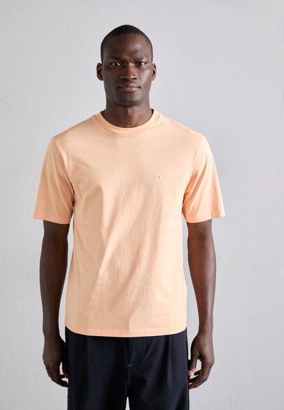 Brzoskwiniowy t-shirt wykonany z bawełny, z okrągłym dekoltem, krótkimi rękawami i małym białym logo na klatce piersiowej, zestawiony z ciemnymi szortami.