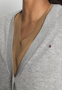 Szary kardigan z głębokim dekoltem w serek, nałożony na brązowy sweter. Zauważalny haftowany detal logo na kardiganie. Miękka tekstura.