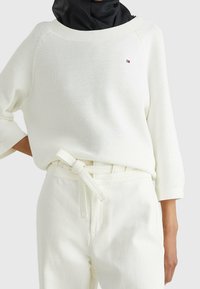 Vit stickad sweatshirt med vid halsringning och trekvartsärmar, kombinerad med högmidjade vita byxor med ett knytbälte.