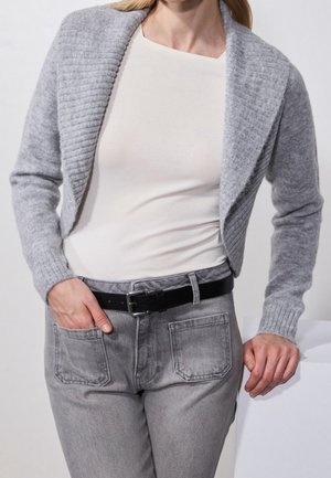 Personne portant un cardigan gris clair court par-dessus un top blanc, jean gris avec une ceinture noire, une main dans la poche, l'autre reposant sur une surface.