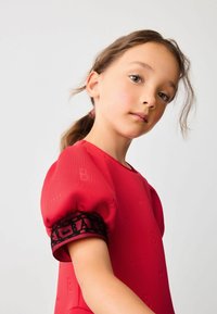 Punainen mekko, joka on valmistettu tekstiilipinnasta ja jossa on puffihihat; se sisältää mustan pitsiraitapannan hihassa, jossa lukee "BAKER."