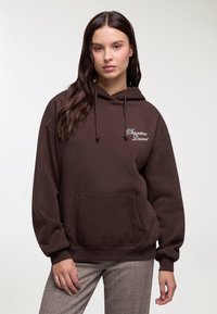 Felpa marrone oversize realizzata in materiale morbido, con tasca frontale, coulisse e testo ricamato bianco. Abbinata a pantaloni a quadri.