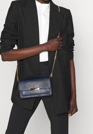 Granatowa torba na ramię z teksturowanym wykończeniem, złotymi okuciami i łańcuszkowym paskiem. Noszona z czarną marynarką na białej bluzce i czarnych spodniach.