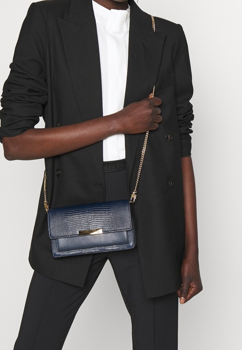 Granatowa torba na ramię z teksturowanym wykończeniem, złotymi okuciami i łańcuszkowym paskiem. Noszona z czarną marynarką na białej bluzce i czarnych spodniach.