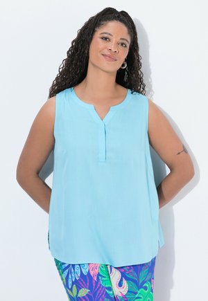 Frau mit lockigem Haar, die ein ärmelloses hellblaues Oberteil und bunte Hosen mit tropischem Muster trägt, steht mit den Händen hinter dem Rücken.