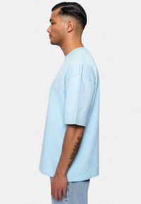 Lyseblå bomulds t-shirt med korte ærmer, afslappet pasform, rund halsudskæring og lille logo-detalje på brystet. Model vist fra siden.