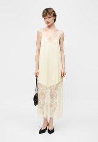 Γυναίκα με αμάνικο φόρεμα σε κρεμ απόχρωση με δαντελωτή τελειώμα, κρατώντας μια μαύρη τσάντα, φορόντας μαύρες μυτερές γόβες, στέκεται μπροστά σε απλό φόντο.