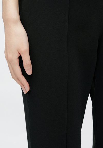 Fekete nadrág sima anyagból, egyenes szabással és finom varrási részlettel az oldalán. A kéz gyengéden nyugszik az anyagon.