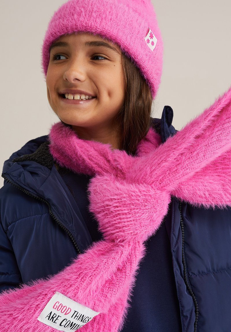 Uśmiechnięta dziewczyna w jasnoróżowej, futrzanej czapce i pasującym szaliku z napisem "Dobre rzeczy nadchodzą", w granatowej kurtce, patrząca na bok.