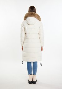 Lang, hvid pufferjakke med en hætte foret med pels, der har en quiltet design og snørebåndsdetaljer i kanten. Båret over blå jeans og støvler.