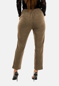 Donna in piedi di spalle che indossa pantaloni di velluto a coste marroni, una maglietta nera trasparente con stampa floreale e scarpe con tacco nere su sfondo bianco.