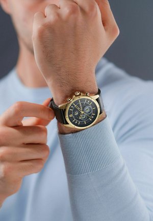 Crna kožna narukvica za sat s kućištem od zlatnog metala, okruglim brojčanikom i sivoj podlozi koja sadrži tri pod-brojčnika i zlatne oznake sati.