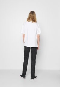 Camiseta de algodón blanca de manga corta, vista desde atrás, combinada con jeans de mezclilla negros y zapatillas de lona negras con suelas blancas.