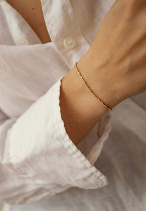 Gold bracelet with a delicate design featuring small, alternating hexagonal and circular links, worn on a wrist with a white linen shirt.