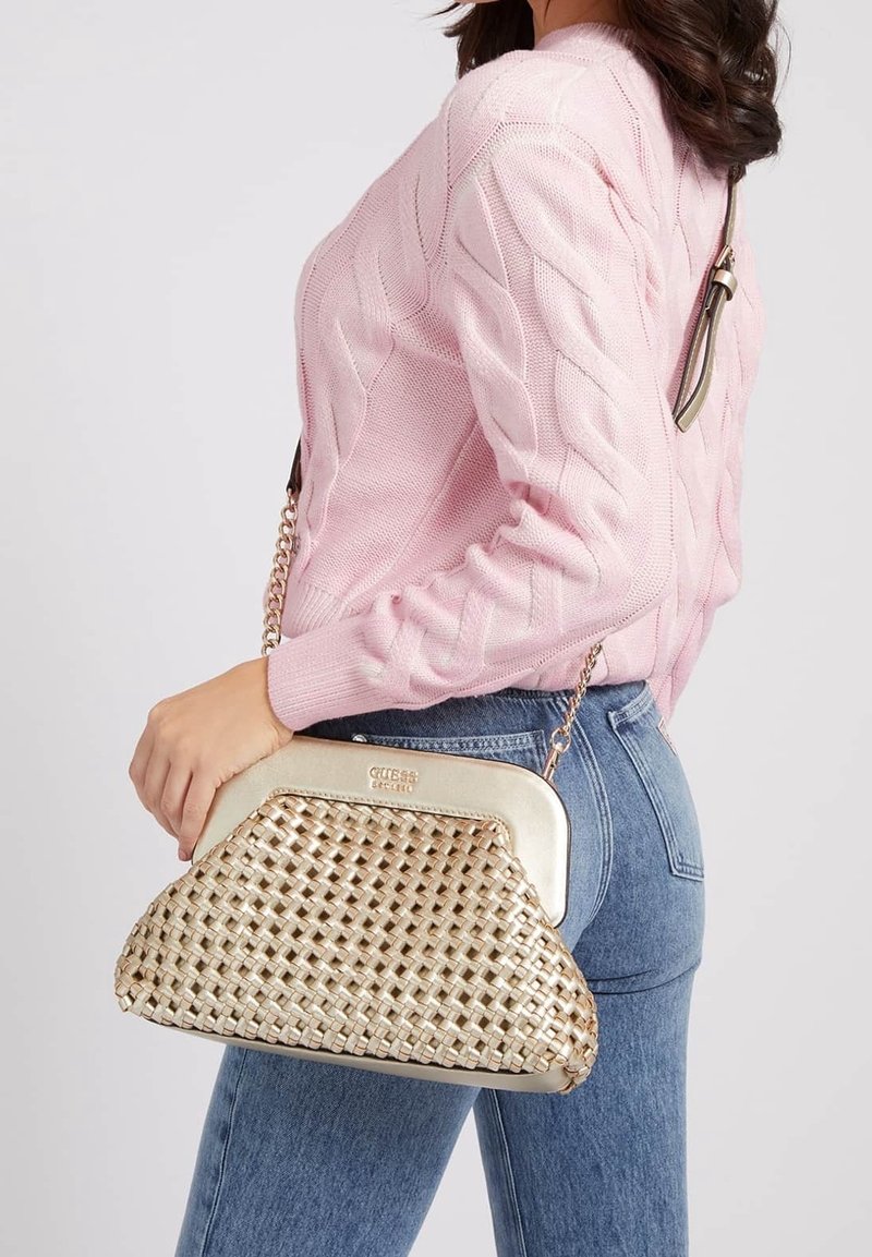 Femme en pull en maille torsadée rose et jean bleu tenant un sac à main beige tissé avec une bandoulière chaîne dorée sur l'épaule.