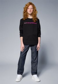 Čierny mikina s ružovým logom "EA7 EMPORIO ARMANI", skombinovaná s tmavomodrými džínsami a bielymi teniskami, na neutrálnom pozadí.