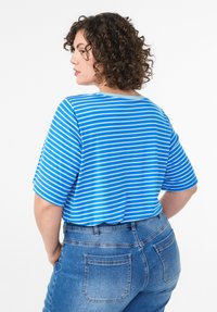 Žena s kučeravými vlasmi, ktorá má na sebe pruhovanú modro-bielu košeľu a modré džínsy, stojí s rukami v vreckách a otočená do strany.