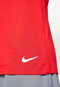 T-shirt de sport rouge à col rond, tissu léger, avec un logo Nike blanc sur le côté bas gauche. Aspect texturé, finition lisse.